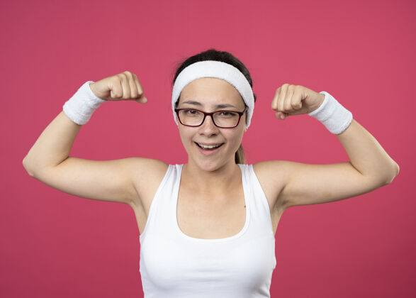 佩戴戴着眼镜 戴着头带和腕带的快乐的年轻运动型女人把二头肌紧绷在粉红色的墙上女人眼镜人