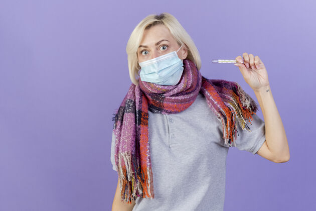 表情一个戴着医用面罩和围巾的年轻金发病女人拿着隔离在紫色墙上的温度计围巾穿着年轻人