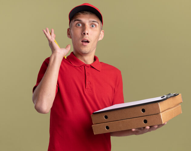 兴奋兴奋的年轻金发送货员举起手站在橄榄绿的比萨饼盒上 手里拿着剪贴板绿色披萨盒子