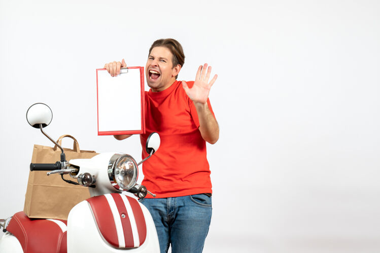 文件俯视图：身穿红色制服 情绪紧张的年轻送货员站在滑板车旁 在白墙上展示文件站立运动微笑