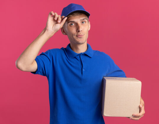 帽子年轻的金发送货员高兴地把手放在帽子上 手里拿着粉色的名片盒年轻请名片盒