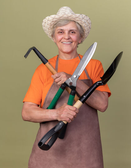 女微笑的上了年纪的女园丁戴着园艺帽手持园艺工具在橄榄绿上园艺花园橄榄
