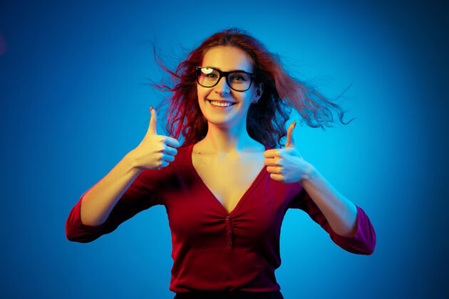 衬衫在霓虹灯下的蓝色工作室背景上孤立的高加索女性肖像休闲风格的红发美女模特人类情感的概念 面部表情 销售 广告竖起大拇指执行女性成功