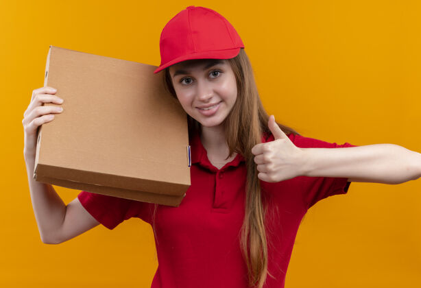 红色身着红色制服 面带微笑的年轻送货员肩上扛着包裹 在孤立的橙色空间竖起大拇指女孩盒子微笑