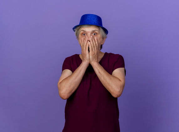 女人震惊的老妇人戴着党的帽子把手放在嘴上孤立的紫色墙壁与复制空间戴上老人帽子