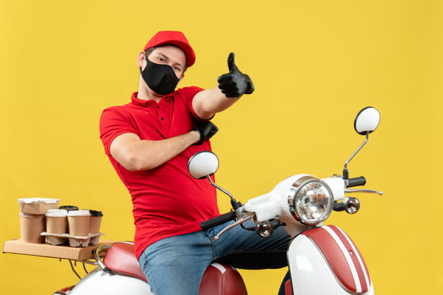 Ok俯视图：微笑的年轻人穿着红色上衣 戴着帽子 戴着手套 戴着医用面罩 坐在滑板车上 在黄色的墙上做着“ok”的手势手套摩托车面罩