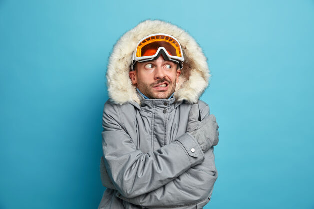 人不满的未刮胡子的年轻人因寒冷而颤抖 他戴着滑雪镜和冬衣拥抱着自己成人快乐摇晃