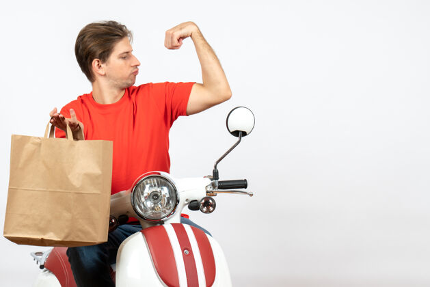 骄傲穿着红色制服的年轻自豪的送货员坐在踏板车上 手里拿着纸袋 在白墙上展示着他的肌肉年轻人滑板车纸