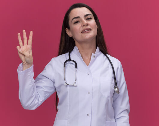 医生高兴的年轻女医生穿着医用长袍 用听诊器做手势 三个手指放在粉红色的衣服上女手指请