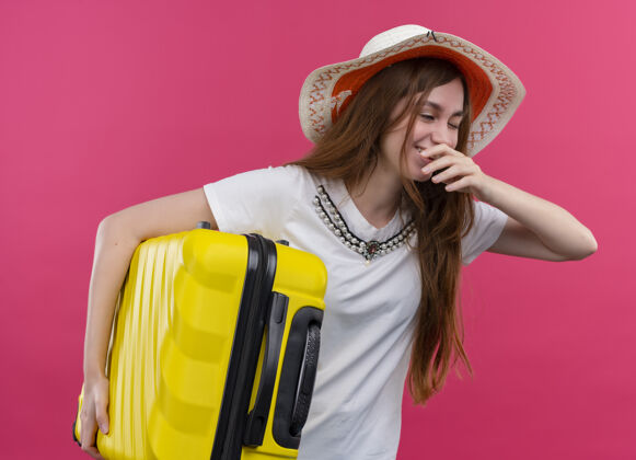 戴着笑着的年轻旅行家女孩戴着帽子拿着手提箱 把手放在嘴边孤立的粉色空间里旅行者帽子手