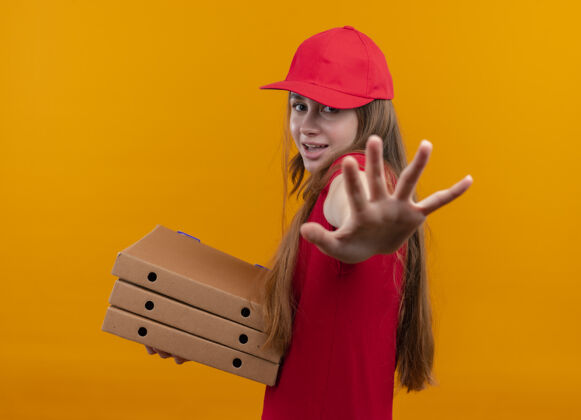 举行快乐的年轻送货女孩穿着红色制服 手持包裹 伸出手 站在孤立的橙色空间的侧视图上橙色交付制服