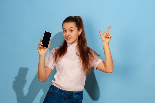 财务展示手机屏幕 指向上蓝色背景上的白人少女肖像穿着休闲服的漂亮模特人类情感的概念 面部表情 销售 广告看起来很开心成功个人教育