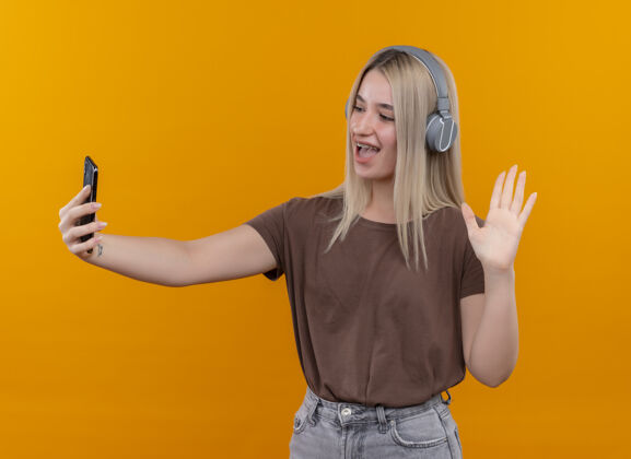 金发快乐的年轻金发女孩戴着耳机戴着牙套拿着手机看着它在孤立的橙色空间挥手手机年轻喜悦
