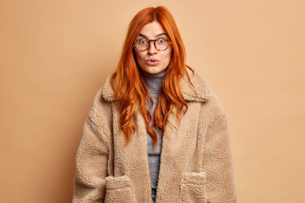 模特惊讶的红发欧洲女人听到难以置信的谣言或秘密印象深刻的东西戴着透明眼镜和皮毛棕色外套成人眼镜情感