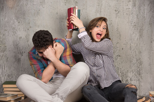 年轻年轻愤怒的女人拿着书打男人可爱锻炼地板