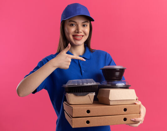 拿着微笑着的漂亮送货员穿着制服 拿着并指着食品包装和容器上的比萨饼盒上的粉红色披萨送货容器