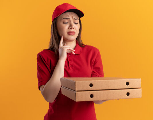 失望一个穿着制服的失望的漂亮送货员把手指放在下巴上 手拿着披萨盒 看着橙色的披萨盒盒子手指下巴
