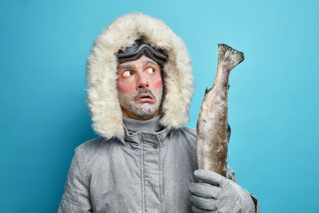 休闲体贴的冻得没刮胡子的男人拿着大奖杯在冰冷的湖面上钓到的鱼在冬天穿着夹克积极休息渔夫护目镜雪