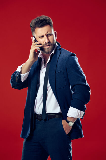 脸帅气的商人拿着手机严肃的商人孤立地站在红色工作室的背景上漂亮的男性半身像人类的情感 面部表情概念信息手机成功