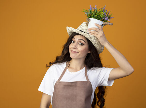 花园穿着制服 戴着园艺帽的年轻女园丁高兴地把花盆举过头顶 隔离在橙色的墙上 留有复制空间拿着园艺制服