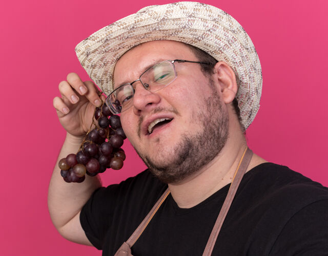 人高兴的年轻男园丁戴着园艺帽 手里拿着葡萄 前面隔离在粉红色的墙上表情年轻人人