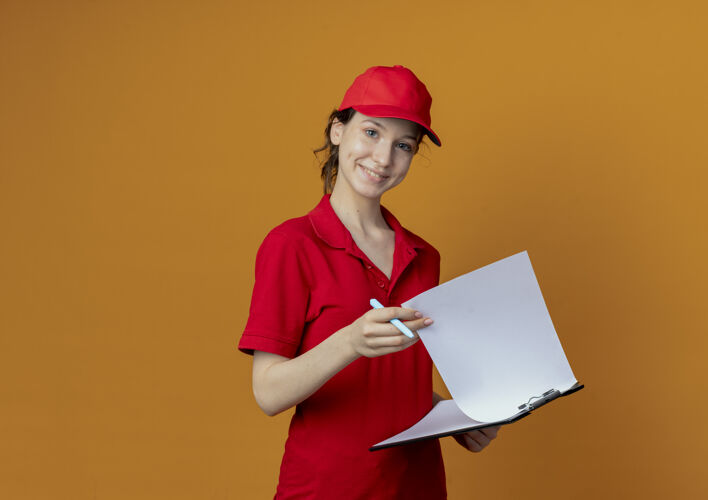 漂亮微笑着的年轻漂亮的送货女孩 穿着红色制服 戴着帽子 拿着剪贴板和笔 孤立地放在橙色背景上 还有复印空间微笑拿着帽子