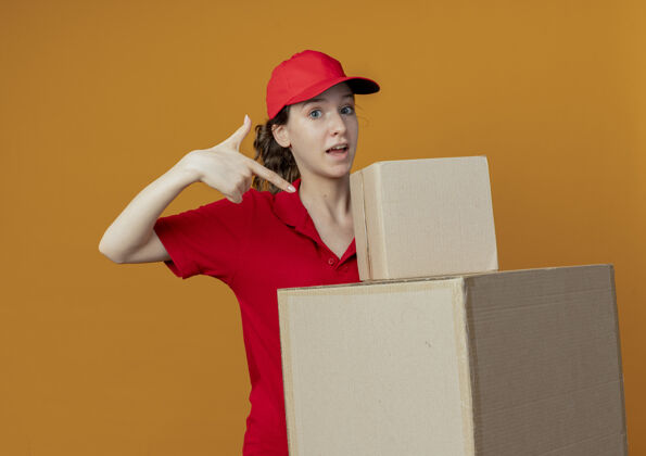 背景令人印象深刻的年轻漂亮的送货女孩在红色制服和帽子举行 并指着纸箱孤立的橙色背景帽子交货橙色