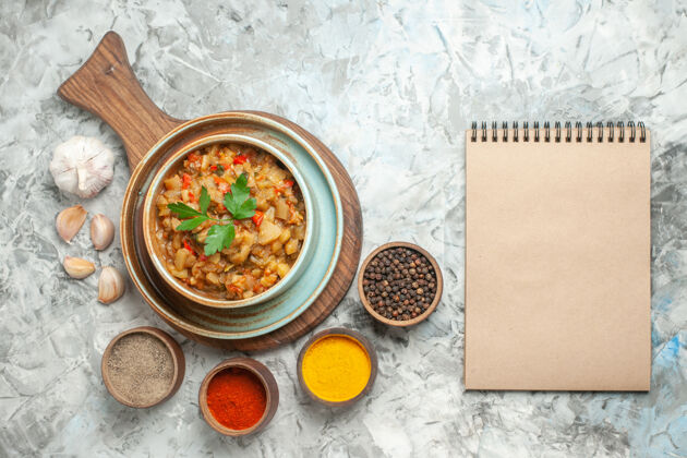 食物砧板上碗里烤茄子沙拉和碗里不同香料的俯视图灰色表面上的笔记本碗板香料