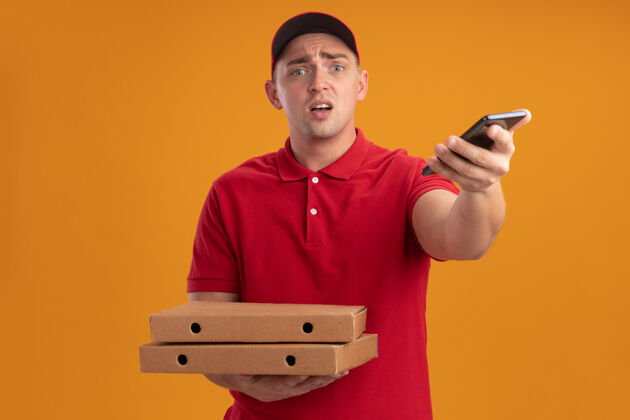 披萨困惑的年轻送货员穿着制服 戴着帽子 手里拿着比萨饼盒 拿着电话站在橙色的墙上电话盒子困惑