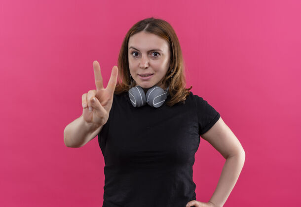 标志自信的年轻休闲女士戴着耳机在脖子上做和平的标志 在孤立的粉色空间年轻粉色脖子