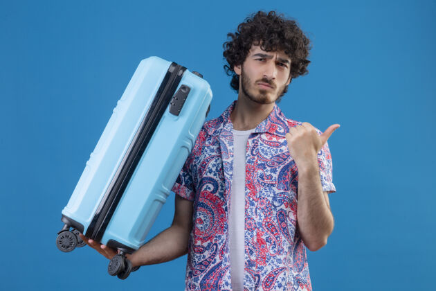 困惑迷茫的年轻英俊的旅行家男子拿着手提箱指着右边的蓝色空间和复印空间指向手提箱帅哥