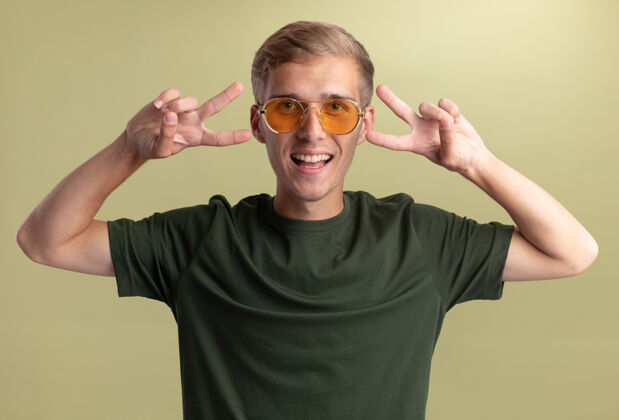 人微笑着的年轻帅哥穿着绿色衬衫 戴着眼镜 在橄榄绿的墙上显示出与世隔绝的和平姿态橄榄表演衬衫