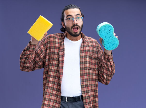 帅哥惊讶的年轻帅气的清洁工穿着t恤拿着海绵隔离在蓝色的墙上惊喜人男人