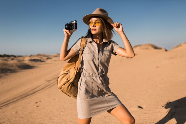 太阳镜穿着卡其色衣服的时髦年轻女子在沙漠中行走 在非洲旅行 戴着帽子和背包 用老式相机拍照自然沙丘体验