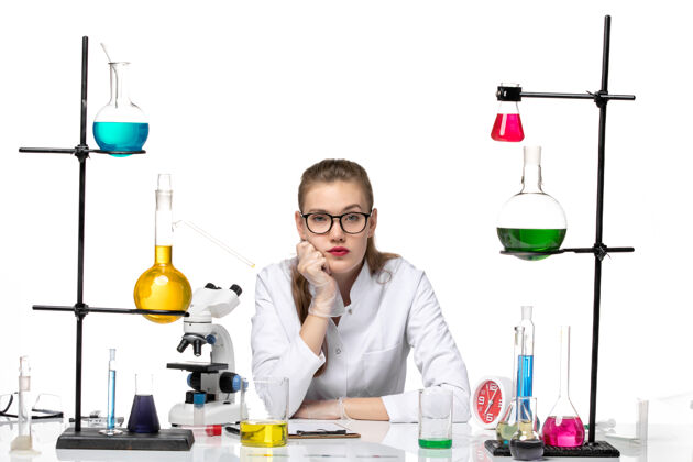 专业前视图：身着医疗服的女化学家在桌子前拿着浅白背景上的溶液大流行化学冠状病毒女化学家前医学