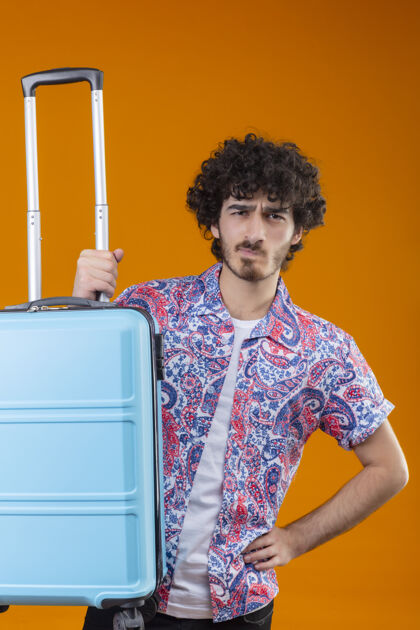 抱着迷茫的年轻英俊卷曲的旅行家男人手放在腰上提着手提箱在孤立的橙色空间年轻手提箱橘子