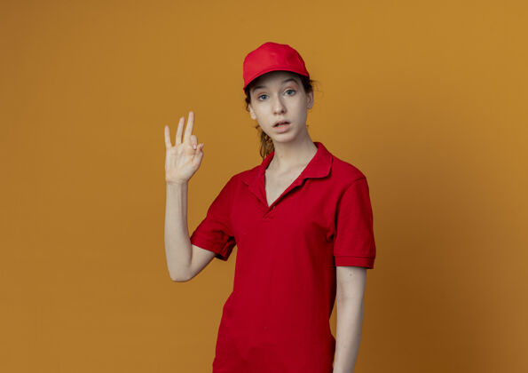 漂亮令人印象深刻的年轻漂亮的送货女孩 穿着红色制服 戴着帽子 做着“ok”的手势 看着隔离在橙色背景上的相机 还有复印空间背景标志帽子