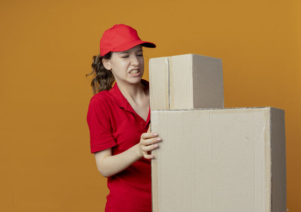 恼火恼怒的年轻漂亮的送货女孩 穿着红色制服 戴着帽子 看着橙色背景上有复印空间的纸箱送货橙色女孩