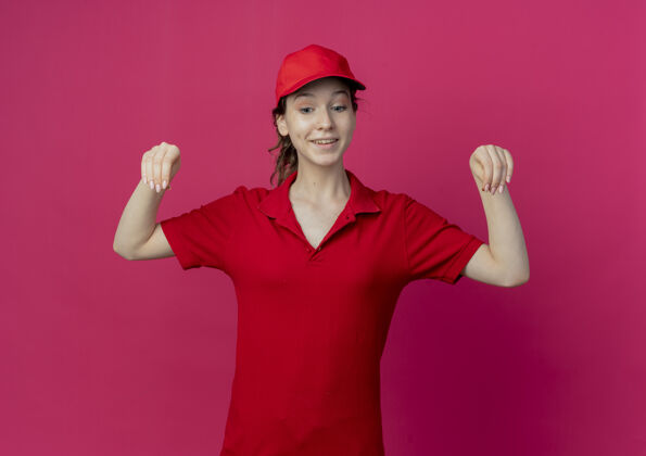 漂亮微笑着的年轻漂亮的送货女孩 穿着红色制服 戴着帽子 低头 假装拿着深红背景上孤立的东西假装制服举行
