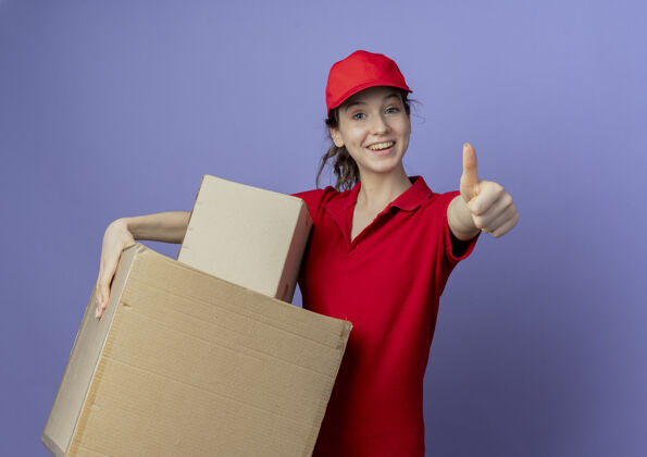 拇指快乐的年轻漂亮的送货女孩穿着红色制服 戴着帽子 拿着纸箱 在紫色背景上的相机前竖起大拇指 留着复印空间美丽年轻欢乐