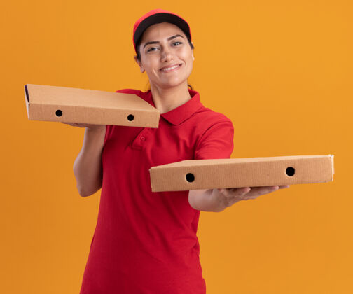 女孩身着制服 头戴鸭舌帽 面带微笑的年轻送货员在橘黄色的墙上举着披萨盒人制服盒子