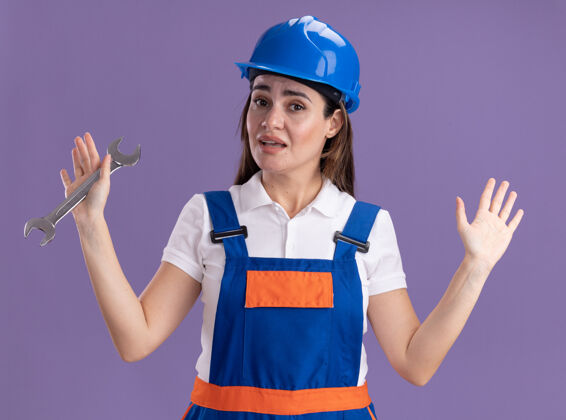 扳手令人印象深刻的年轻建筑妇女在统一举行开放式扳手展开双手孤立在紫色的墙壁脸衣服姿势