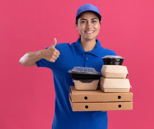 穿着面带微笑的年轻送货女孩 穿着制服 戴着帽子 拿着比萨饼盒上的食物容器 在粉红色的墙上孤立地竖起大拇指女孩脸制服
