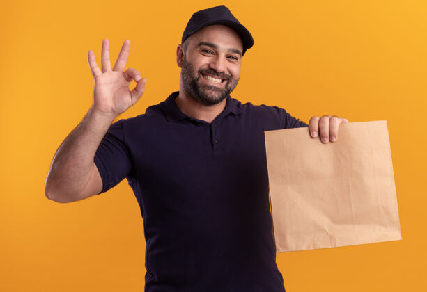 手势微笑的中年送货员穿着制服 戴着帽子 拿着纸食品包 在黄色的墙上显示出良好的姿态食物表情人