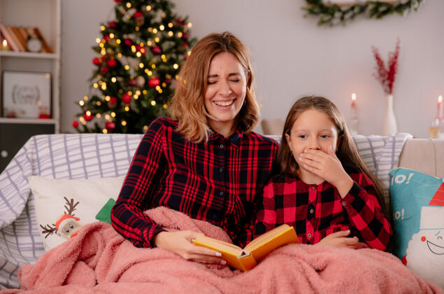 时间笑眯眯的母女俩坐在沙发上读着铺着毛毯的书 在家里享受圣诞节时光坐着房子沙发