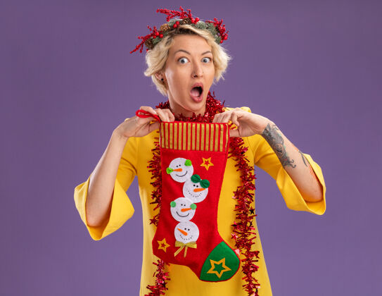 金发年轻的金发女郎戴着圣诞花环 脖子上戴着金属丝花环 手里拿着圣诞长袜 看着紫色背景上孤立的摄像机金属丝圣诞节周围