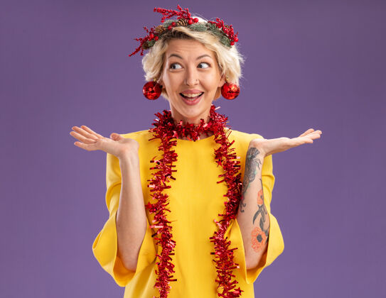 女人令人印象深刻的年轻金发女子头戴圣诞花环和镀金花环在脖子上看一边显示空手圣诞饰品挂在她的耳朵孤立的紫色背景壁板头花环