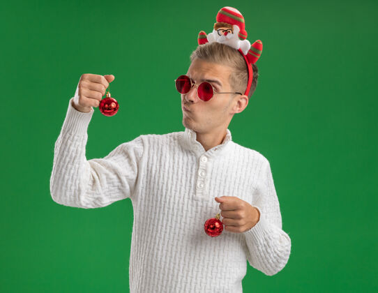 圣诞老人可疑的年轻帅哥戴着圣诞老人的头带 戴着眼镜 手里拿着圣诞饰品 看着其中一个孤立在绿色背景上的圣诞饰品眼镜圣诞老人头带