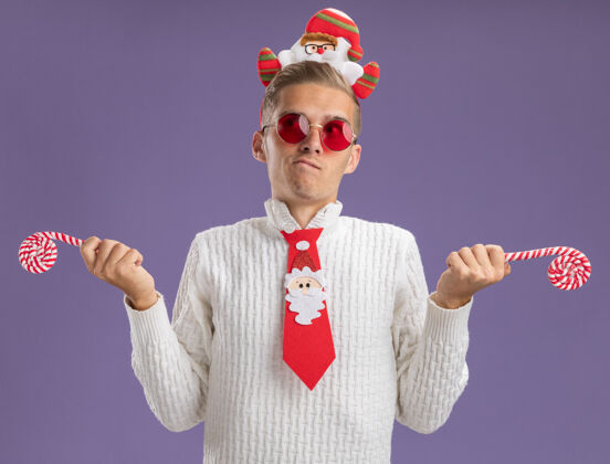 圣诞老人迷茫的年轻帅哥戴着圣诞老人的头带和领带戴着眼镜拿着圣诞糖果手杖看着紫色背景上孤立的一面年轻手杖领带