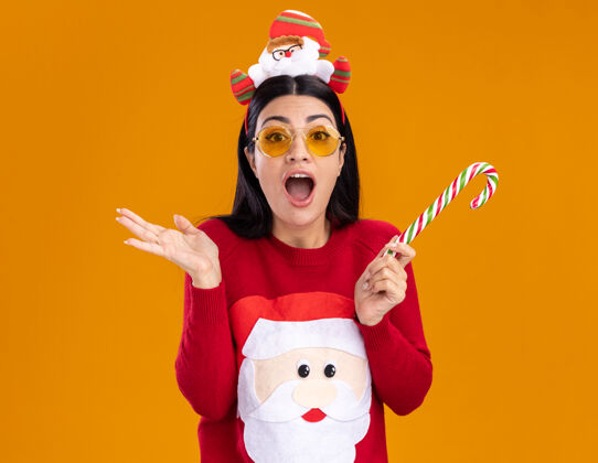 表演令人印象深刻的年轻白人女孩戴着圣诞老人的头带和毛衣 戴着眼镜 手持传统的圣诞糖果手杖 看着相机 在橙色的背景上显示孤立的空手眼镜印象手